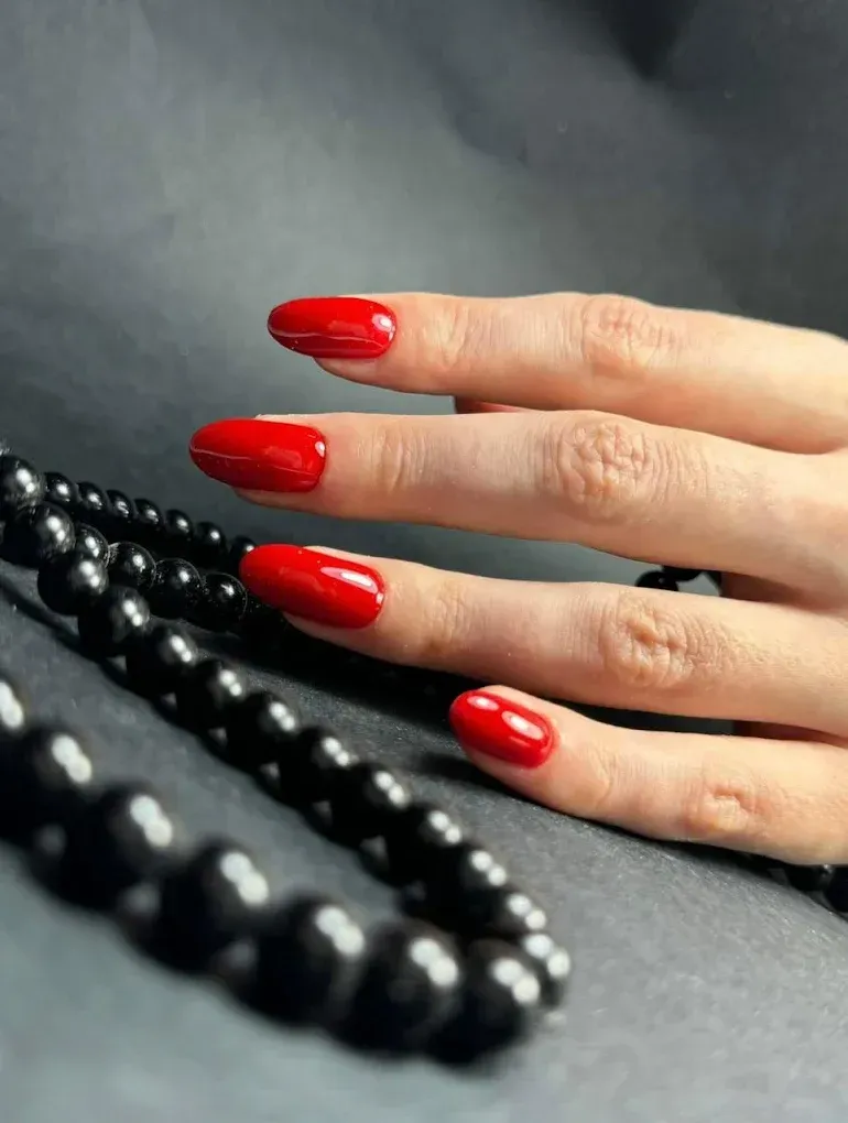 Una mano con uñas rojas brillantes y bien cuidadas descansa cerca de un collar de cuentas negras pulidas sobre un fondo oscuro.
