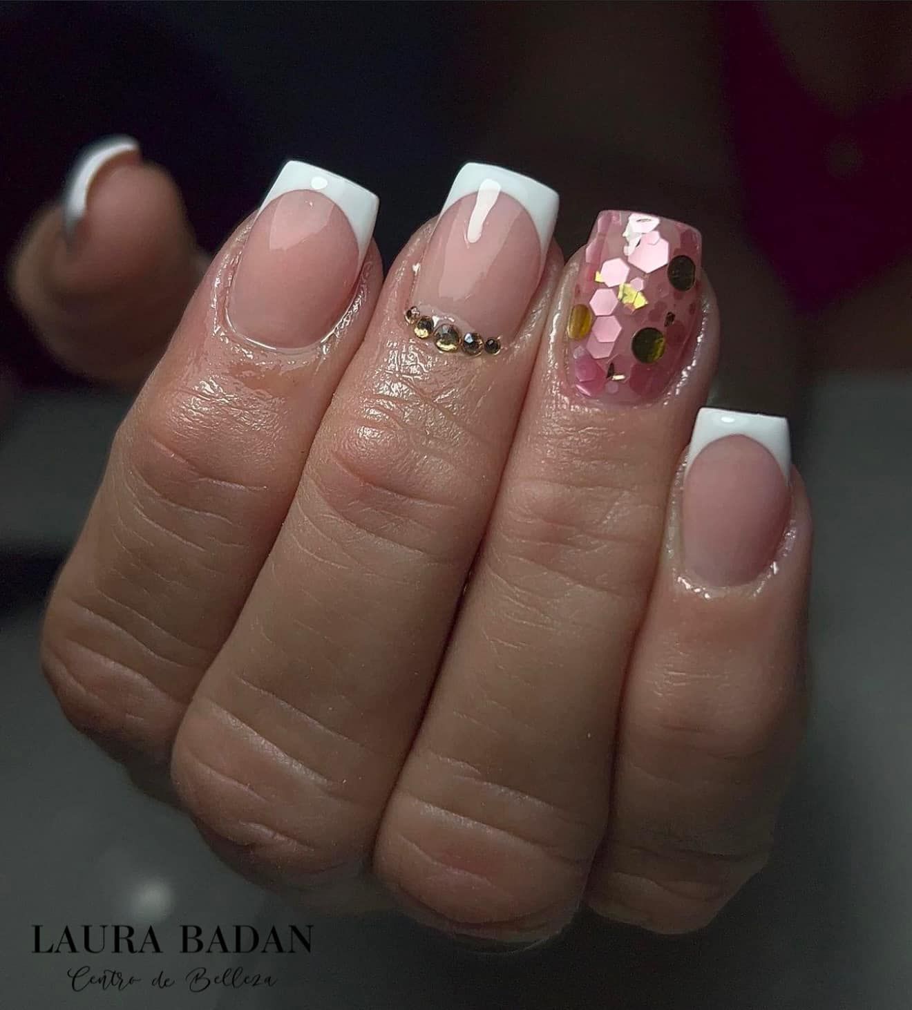 Una mano con uñas de punta cuadrada, de base rosa y puntas blancas, una de ellas adornada con purpurina dorada.