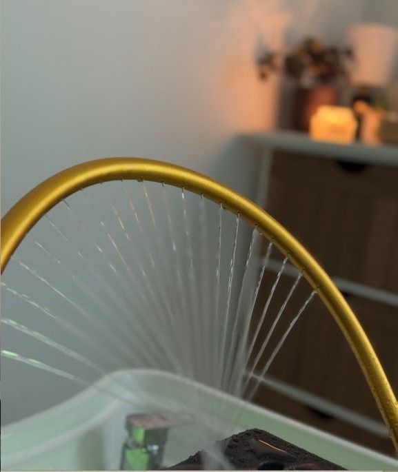 Una lámpara arqueada de color dorado que rocía un chorro de agua en forma de abanico sobre un lavabo blanco, con un entorno interior difuminado al fondo.