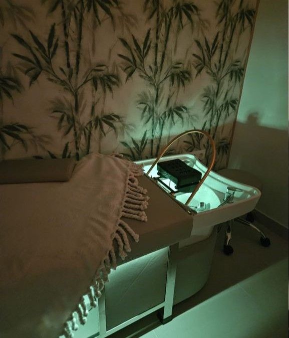 Una sala de peluquería con una camilla de masaje con una manta con flecos, junto a un lavacabezas bajo una suave iluminación ambiental verde.