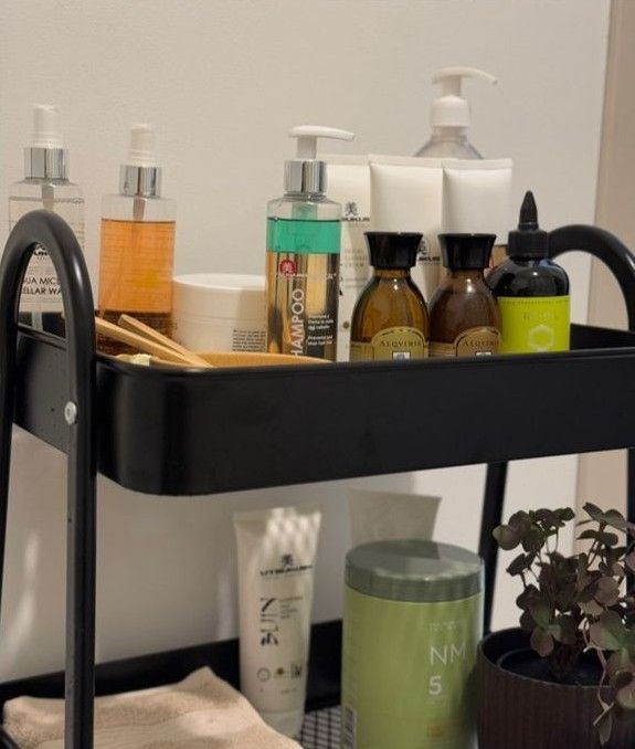 Un carrito rodante de metal negro de dos niveles que contiene diversos productos para el cuidado de la piel y el cabello, con una pequeña planta en maceta en la parte inferior.