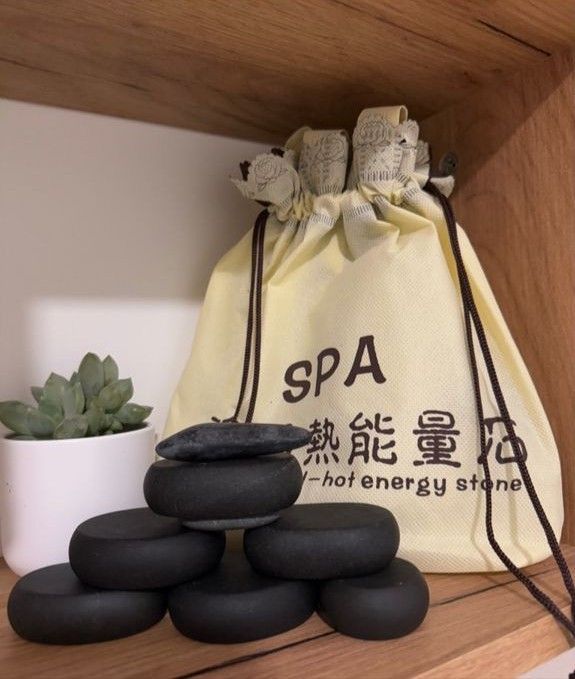 Una pequeña planta suculenta en una maceta blanca y una pila de piedras de spa negras y lisas delante de una bolsa beige con cordón.