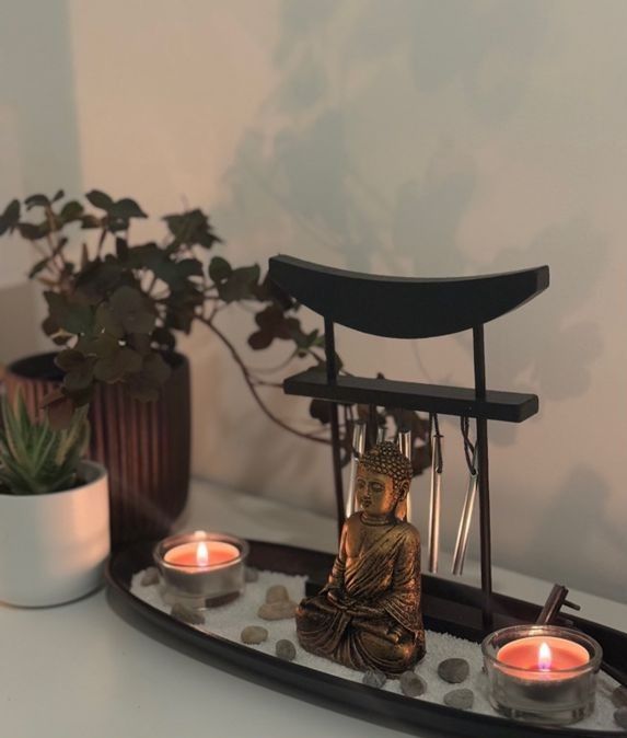 Un jardín zen con una estatua de Buda, dos velas pequeñas encendidas y un marco de puerta torii en miniatura sobre una bandeja.