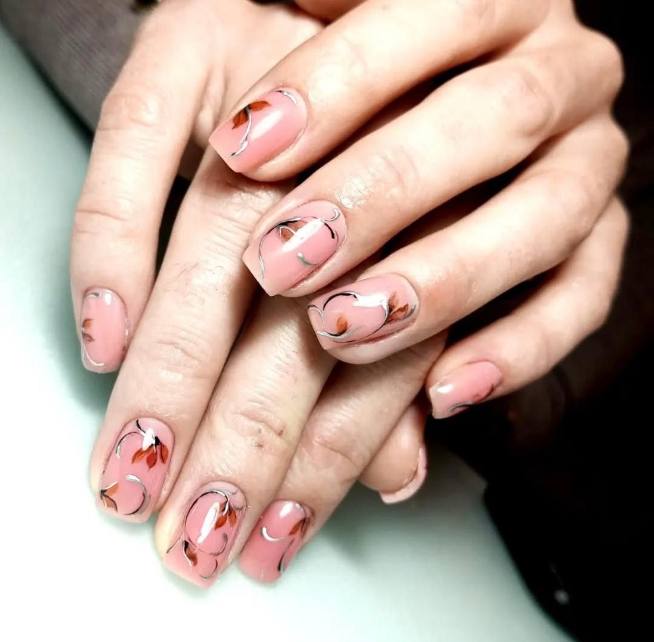 Primer plano de unas manos con una manicura rosa claro adornada con delicados diseños florales en tonos marrones y plateados.