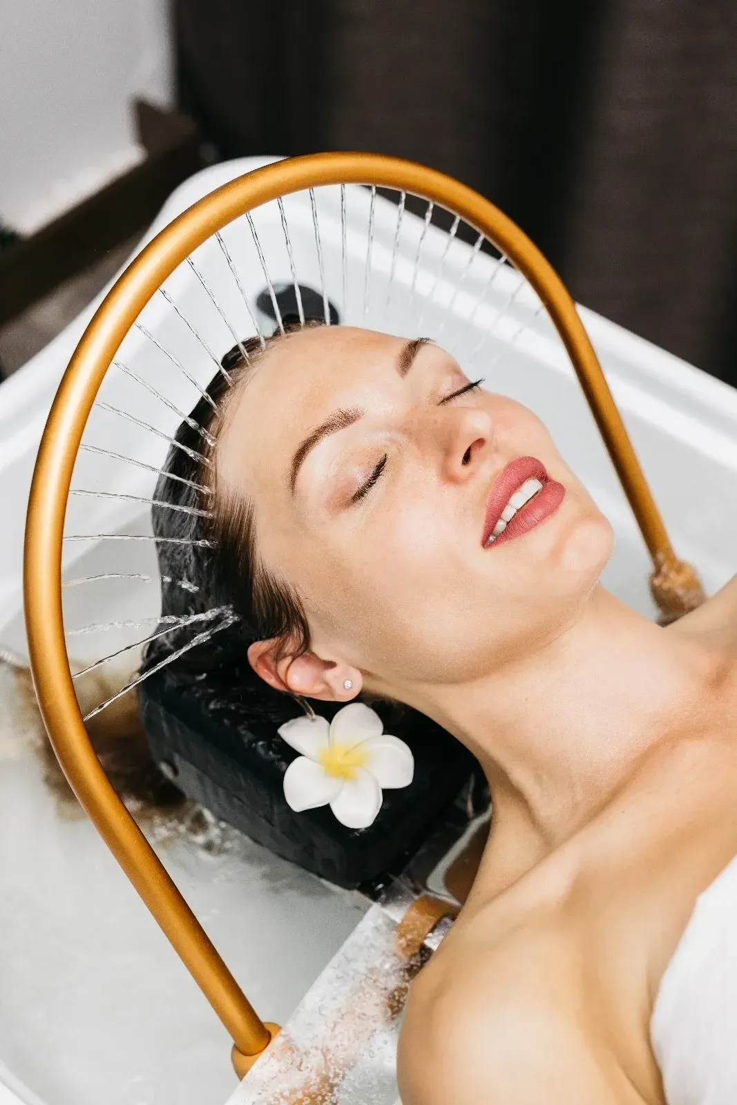 Una persona recostada en una bañera de hidromasaje recibiendo un tratamiento capilar con agua proveniente de una ducha dorada con forma de arco.