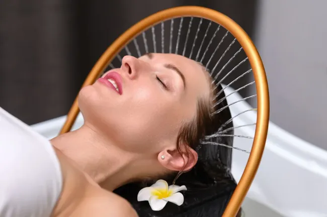 Una persona se relaja con los ojos cerrados mientras una fuente circular rocía agua suavemente sobre su cabello en un ambiente de spa.