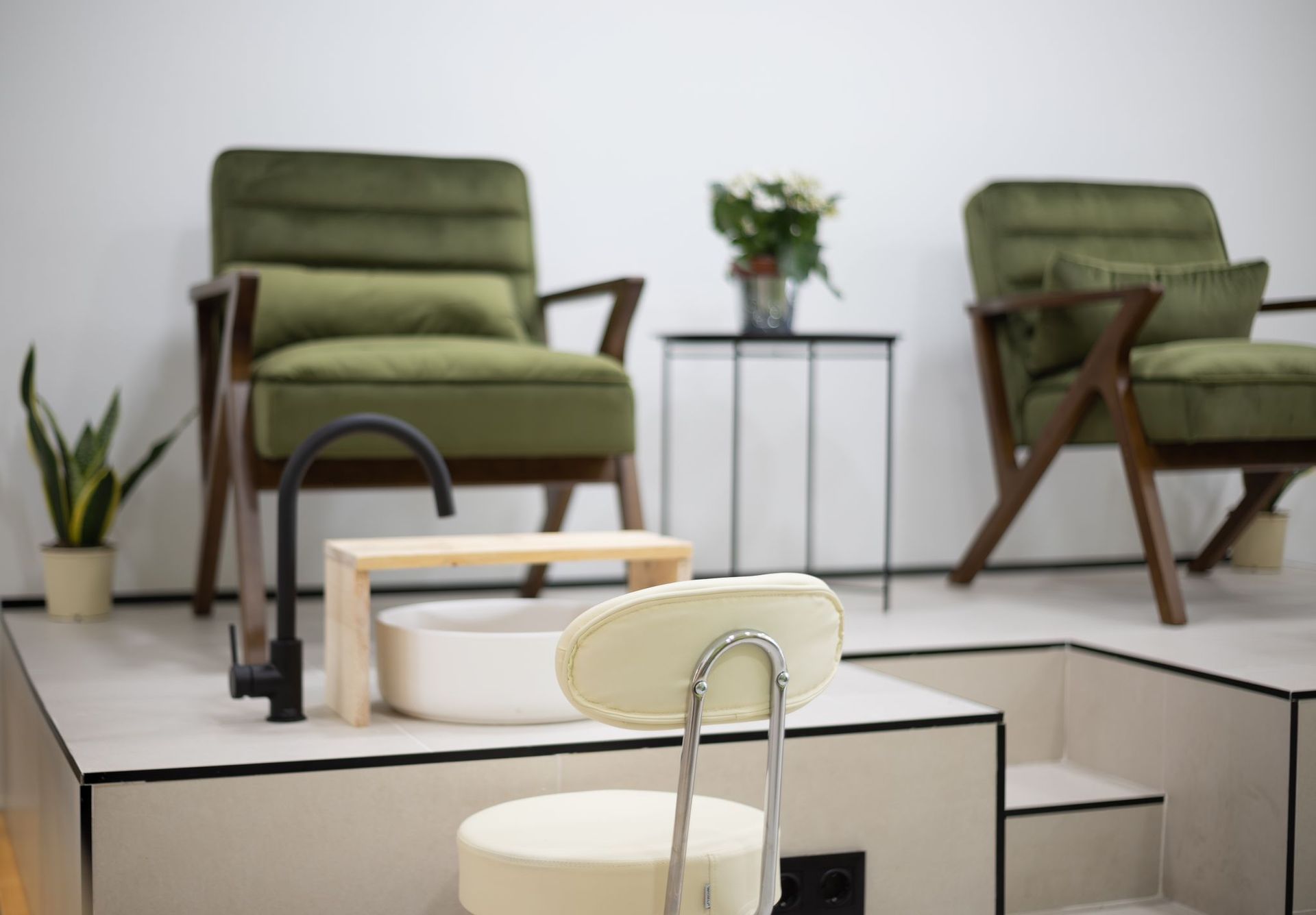 Un puesto de pedicura con lavabo blanco y grifo negro frente a dos sillones de terciopelo verde en un salón minimalista.