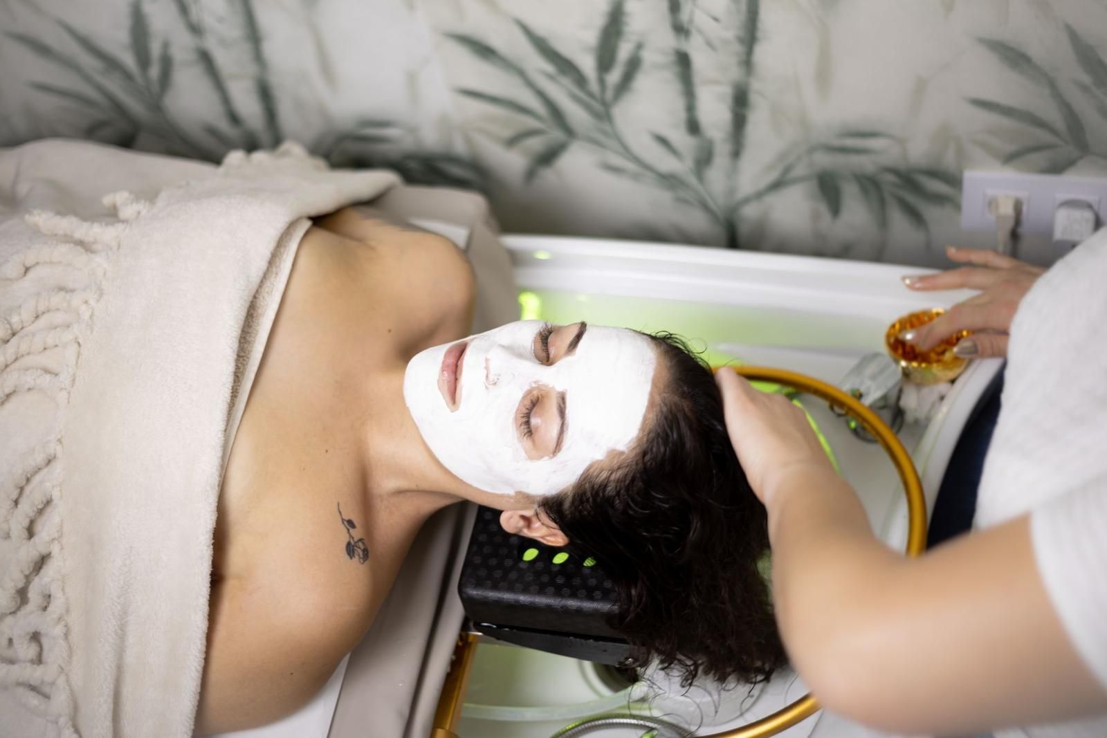 Una persona con una mascarilla blanca se recuesta en el lavabo de una peluquería mientras un profesional le lava el pelo.