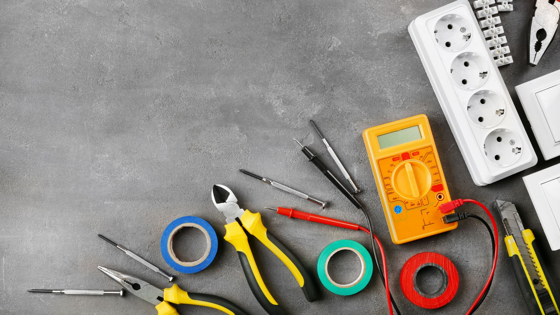 Outils et composants électriques sur une surface grise : multimètre, pinces, ruban adhésif, prises.