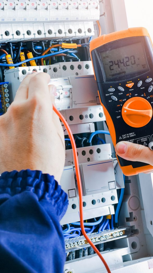 Électricien utilisant un multimètre pour tester le câblage à l'intérieur d'un panneau électrique, portant des manches bleues.