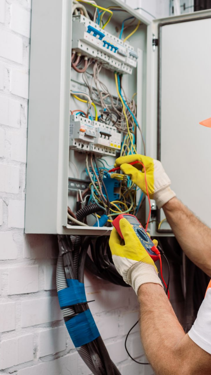 Un électricien portant des gants jaunes teste les fils dans un boîtier de disjoncteur sur un mur de briques.