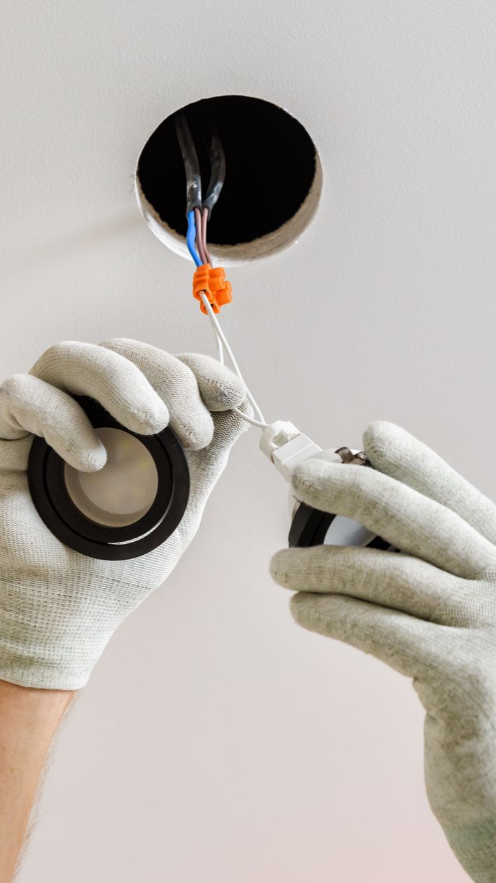 Mains portant des gants installant un luminaire encastré, fils visibles dans le trou du plafond.