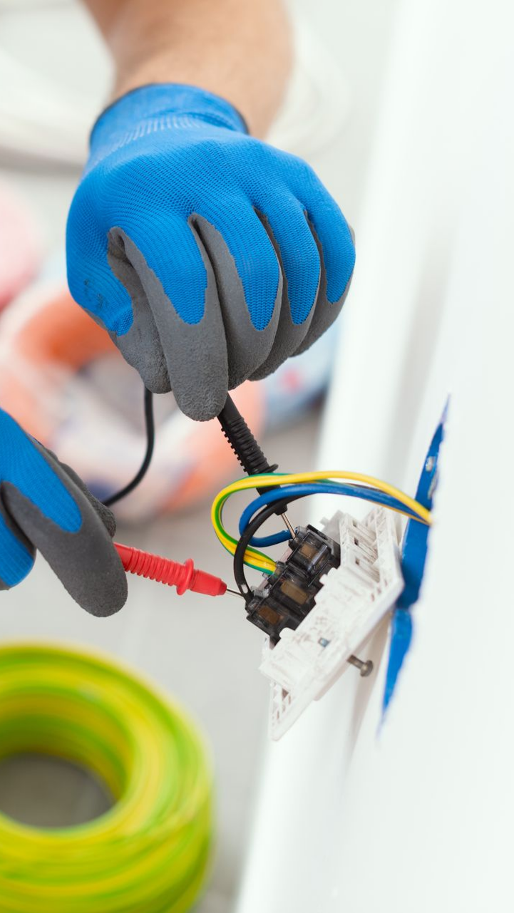 Électricien en gants bleus testant une prise électrique avec des fils ; fil jaune et vert en arrière-plan.