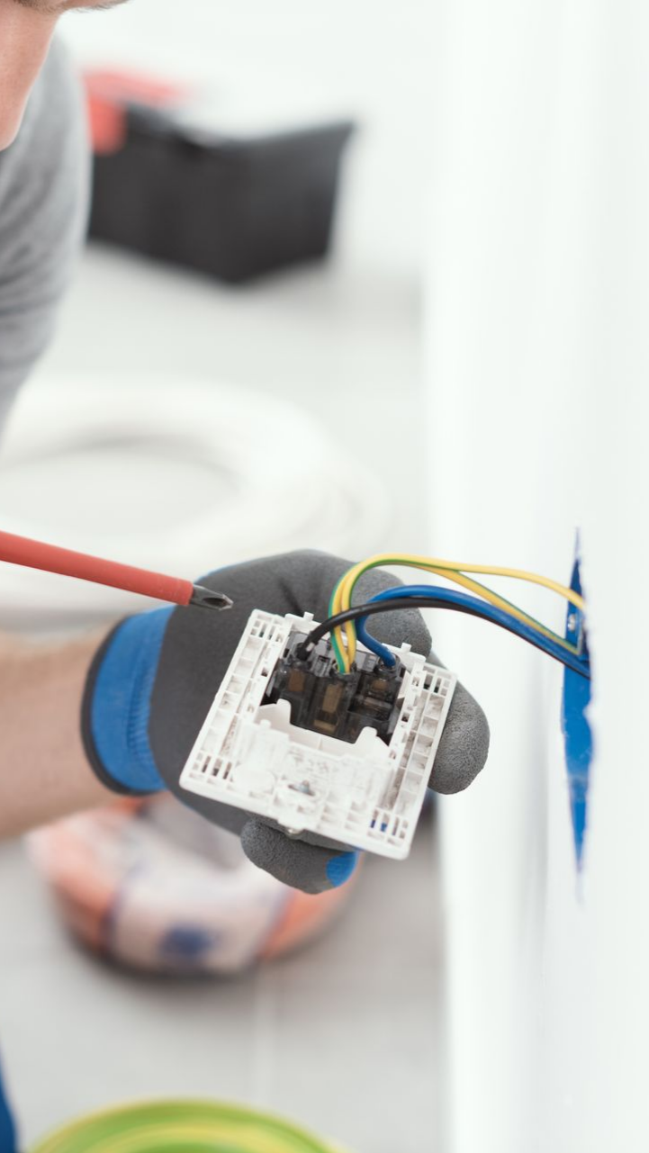 Électricien installant une prise murale, la tenant avec des mains gantées, tournevis en cours d'utilisation.