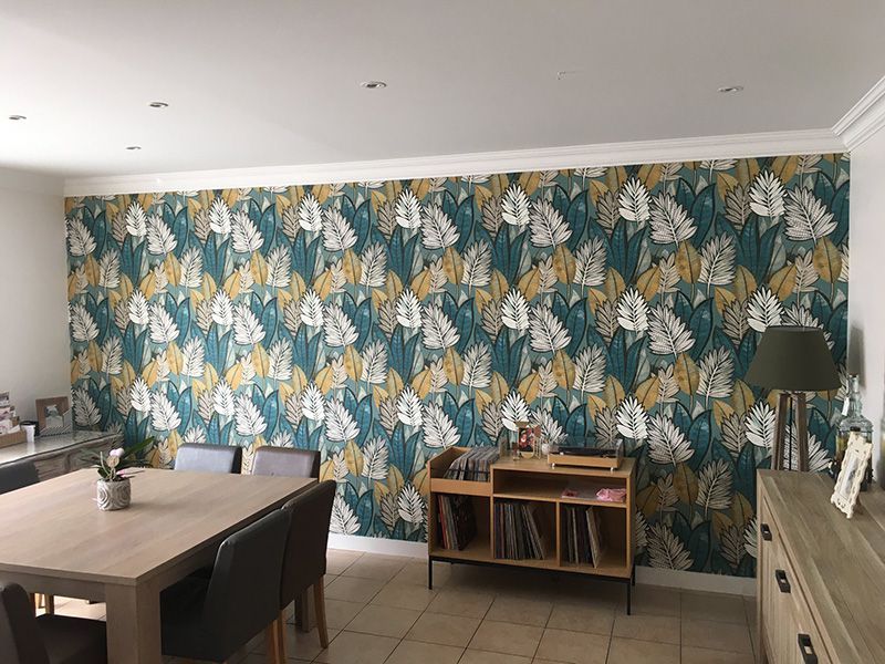 Salle à manger avec papier peint à feuilles bleu sarcelle, or et blanc, table et rangement en bois, meubles neutres.