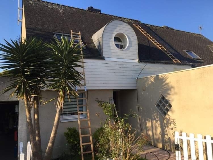 Extérieur de la maison avec lucarne, échelle sur le toit, palmiers et clôture blanche.