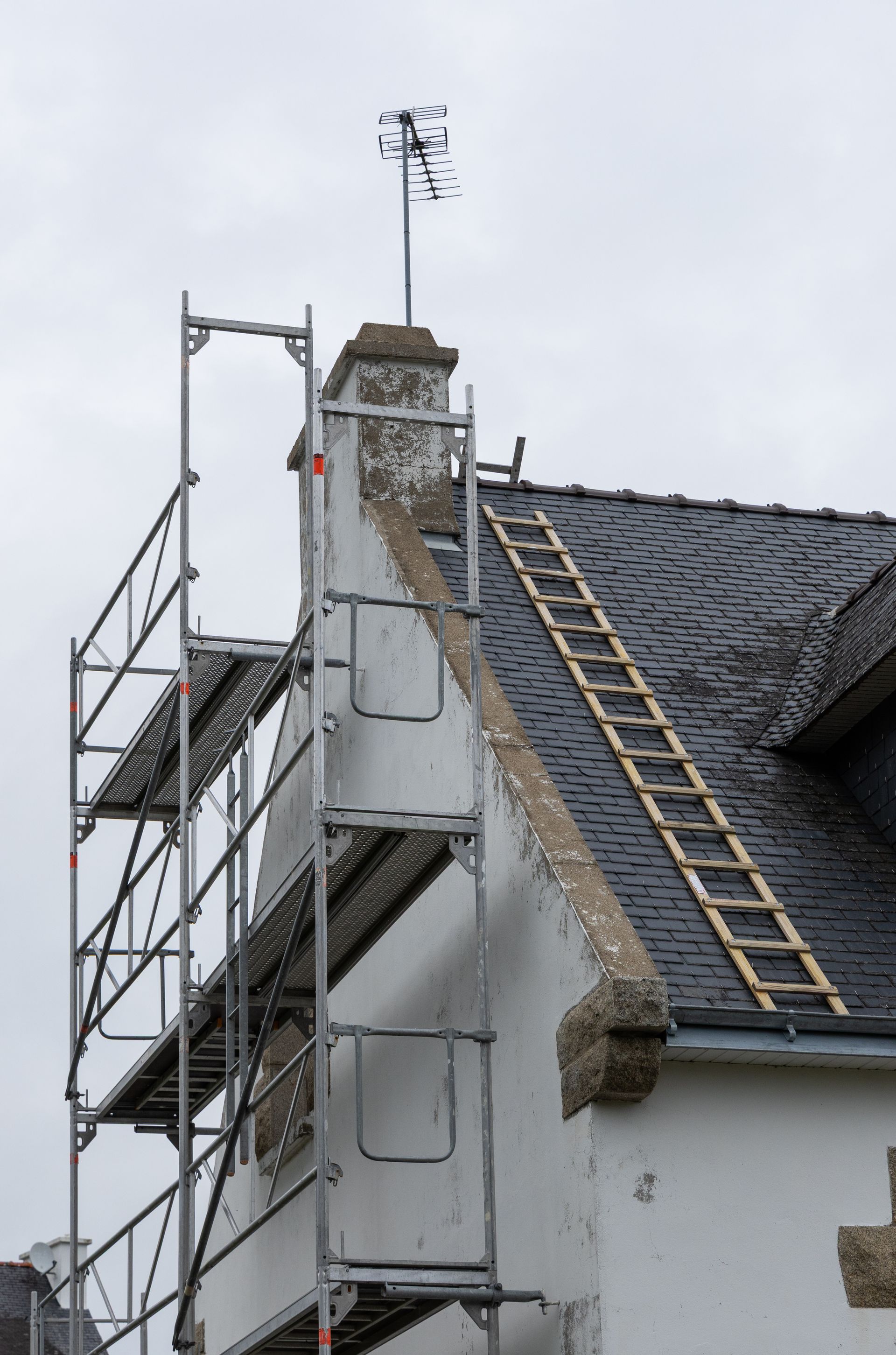 Échafaudage à côté d'une maison blanche avec un toit de tuiles noires et une échelle sur le toit.