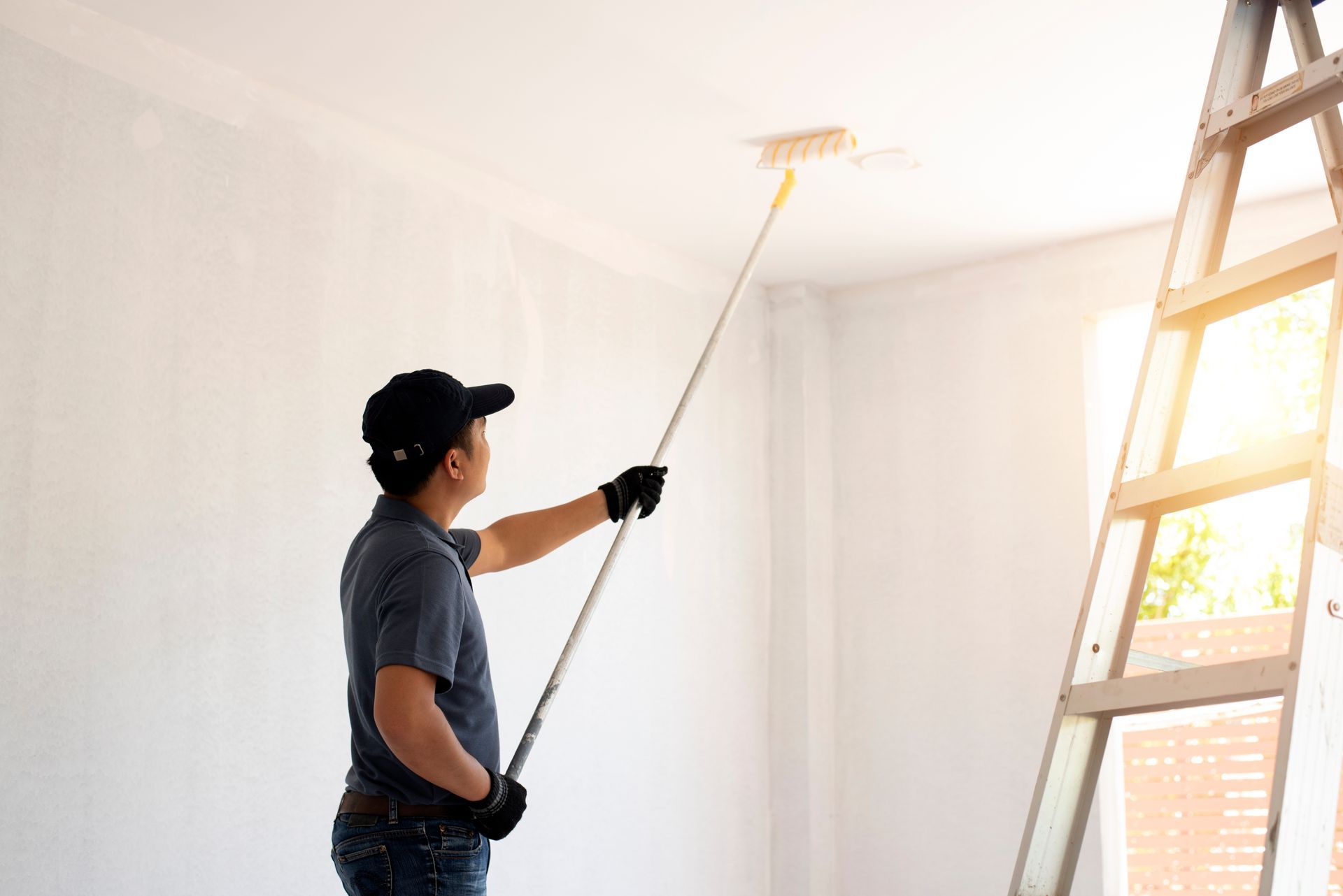 Peintre sur une échelle, peignant un plafond blanc avec un rouleau à long manche dans une pièce bien éclairée.