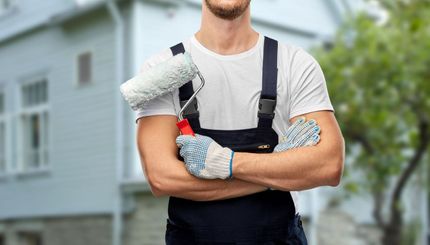 Peintre avec rouleau et gants, souriant, devant une maison.