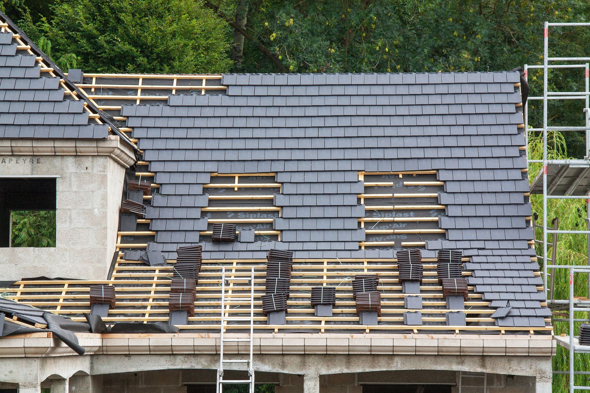 Toiture d'un bâtiment en construction ; tuiles gris foncé en cours de pose sur une ossature bois, avec échafaudage visible.