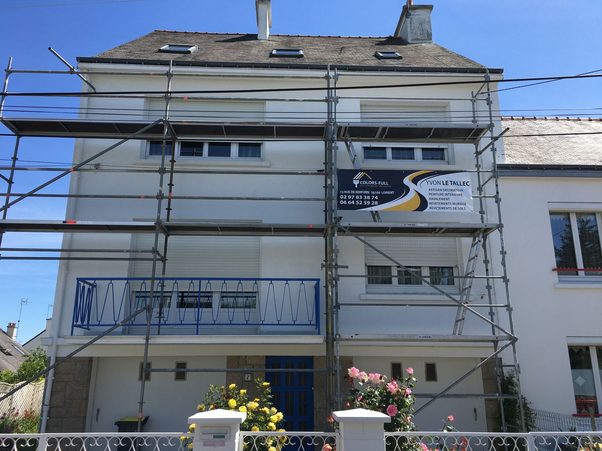 Échafaudage sur une maison à deux étages en cours de peinture blanche. Un panneau avec du texte est visible.