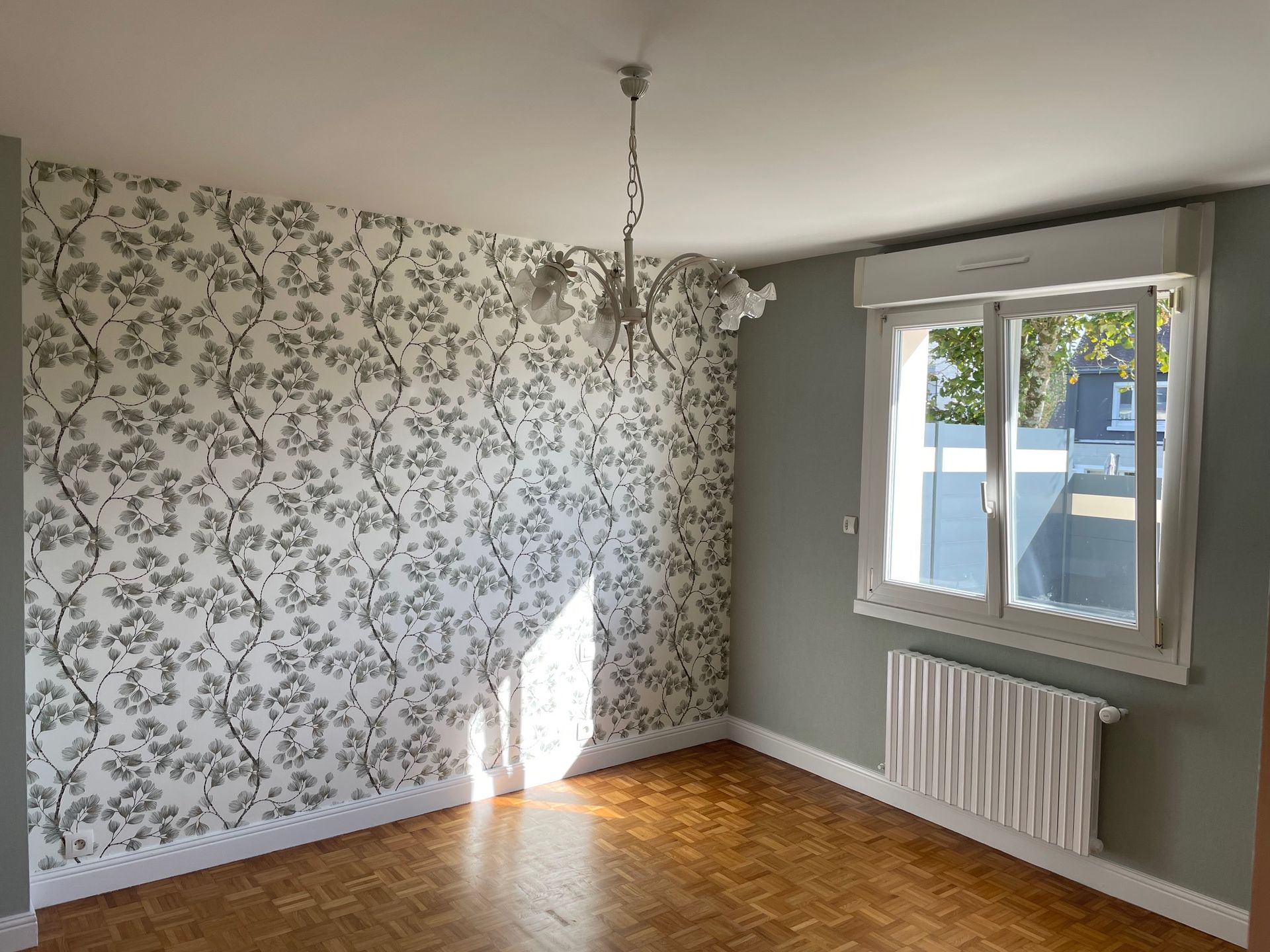 Chambre vide avec papier peint floral, parquet, radiateur et fenêtre.