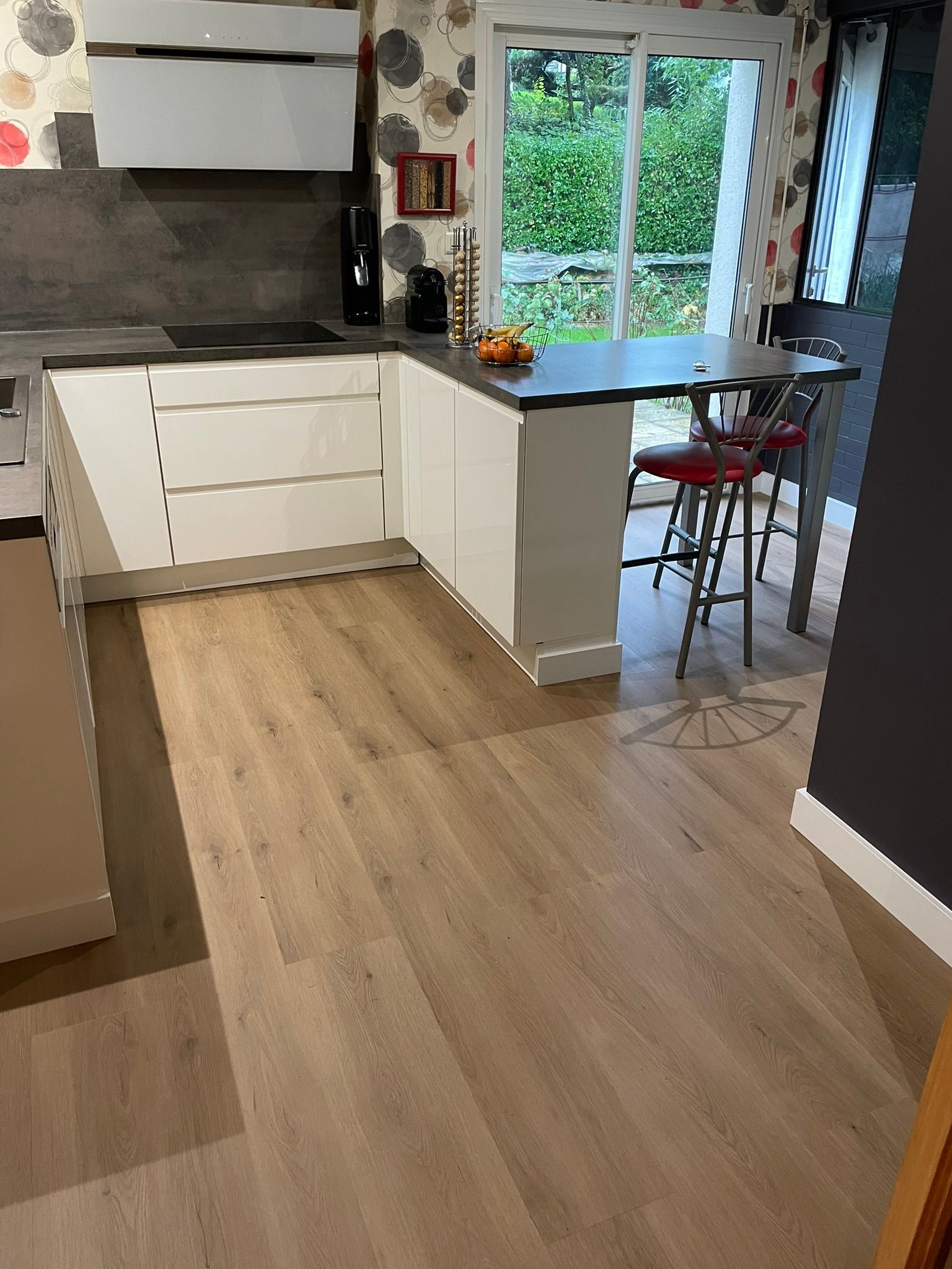 Cuisine avec armoires blanches, plans de travail foncés et parquet. Un bar à petit-déjeuner avec tabourets rouges est installé devant une fenêtre.