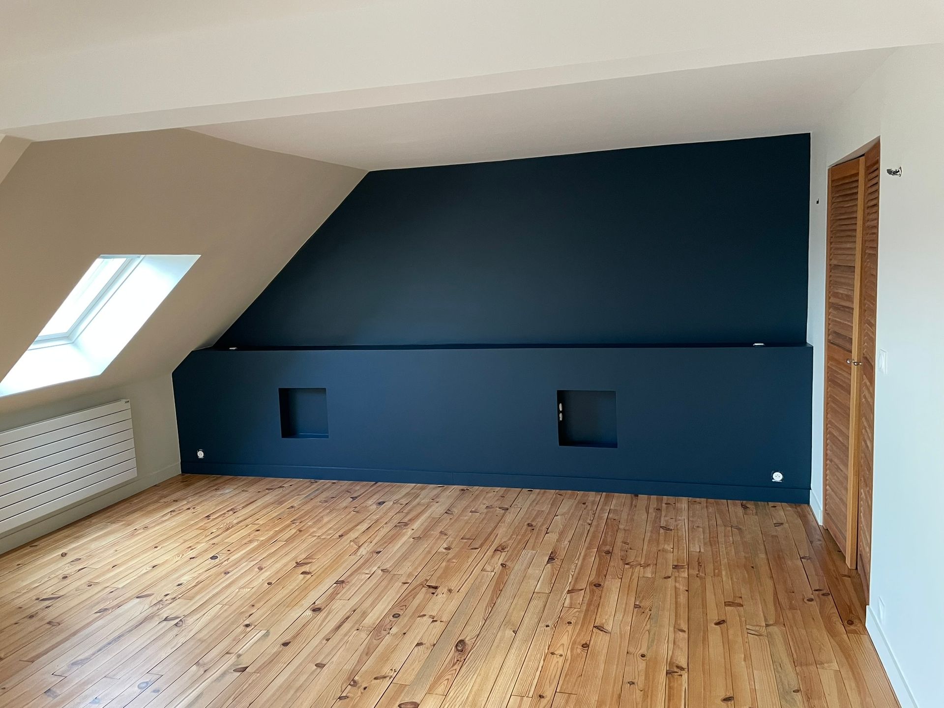 Chambre mansardée avec mur d'accent bleu foncé, plancher en bois, puits de lumière et porte en bois fermée.