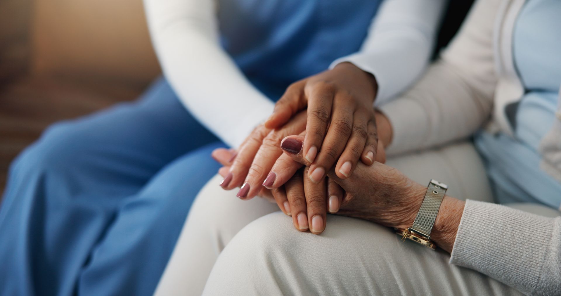 Les mains jointes dans un geste de soutien sur les genoux d’une personne assise, avec des vêtements bleus et beiges visibles.