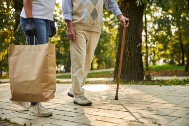 Deux personnes se promènent sur une allée ensoleillée d’un parc, l’une portant un grand sac de courses en papier brun et l’autre s’aidant d’une canne