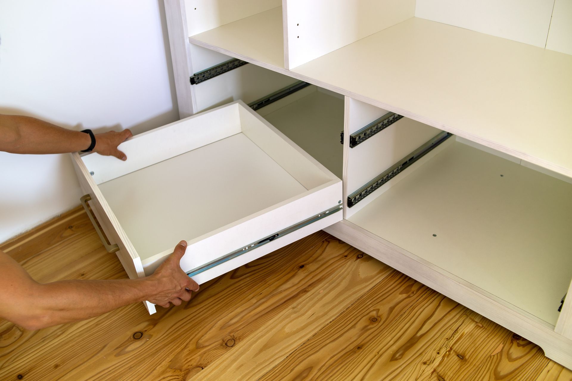 Des mains tirent un tiroir blanc d’un meuble beige avec des étagères, sur un sol en bois