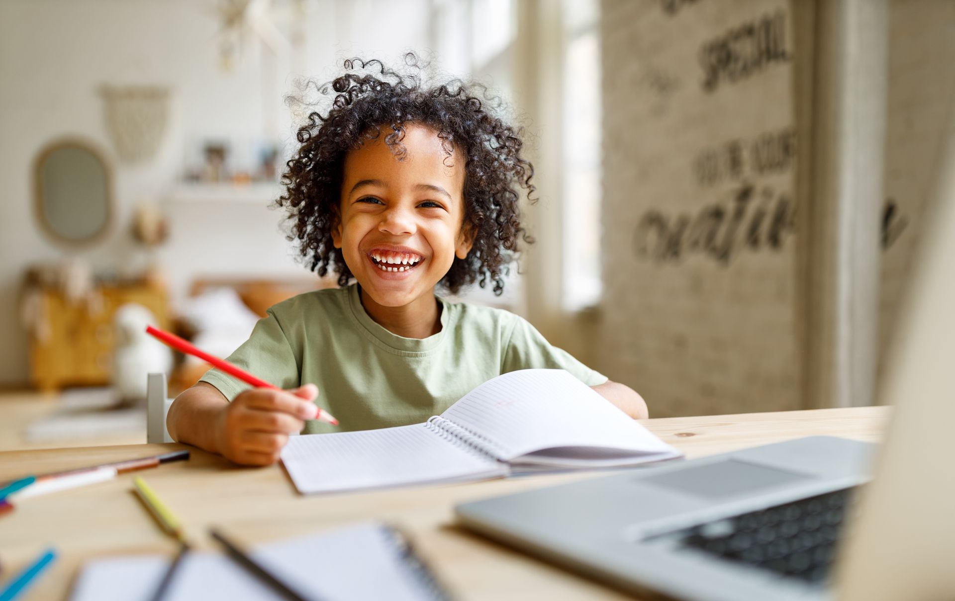 Enfant souriant qui écrit avec un crayon rouge à un bureau, à côté d’un ordinateur portable ouvert et d’un cahier