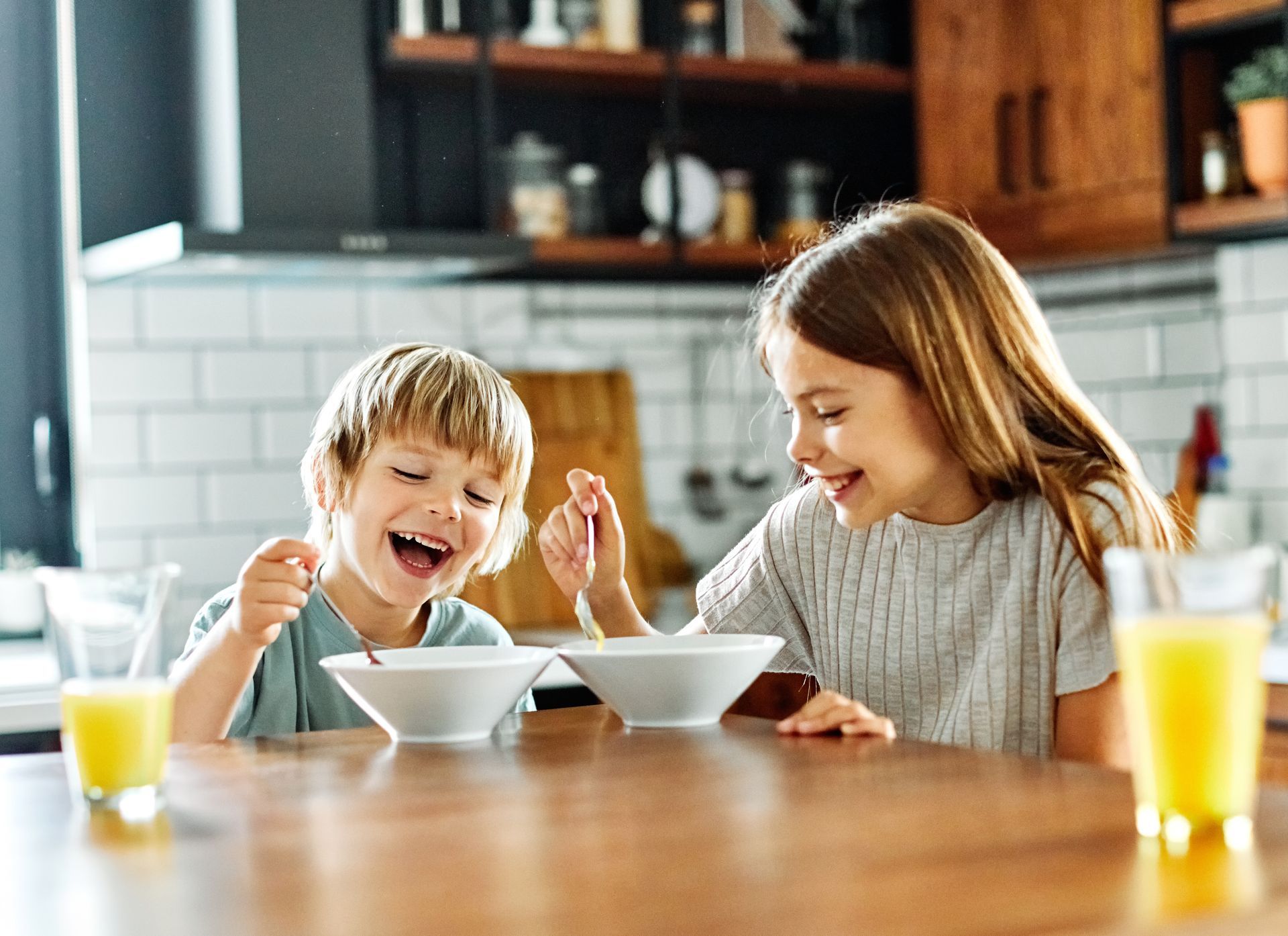 Deux enfants prennent leur petit-déjeuner autour de la table de la cuisine, avec des céréales et du jus d’orange, le sourire aux lèvres