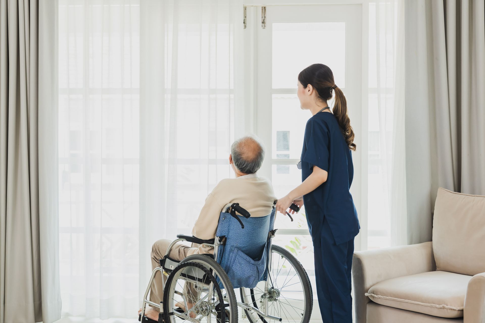 Un aidant aide une personne en fauteuil roulant près d’une fenêtre lumineuse, dans le salon d’une maison