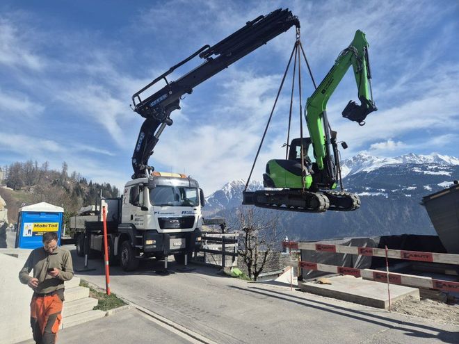 Ein LKW-Kran hebt einen grünen Bagger auf einer Strasse mit Bergblick an. Ein Arbeiter steht daneben.
