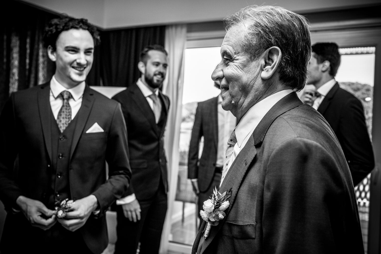 Hombres trajeados conversan, posiblemente antes de una boda, en un interior cerca de una ventana. Un hombre mayor sonríe.