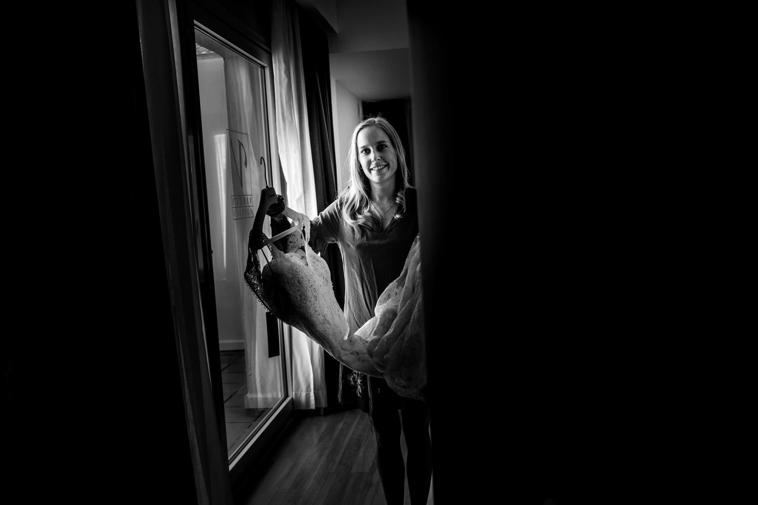 Una mujer sonríe en una puerta, sosteniendo un gran animal de peluche frente a una ventana con cortinas.