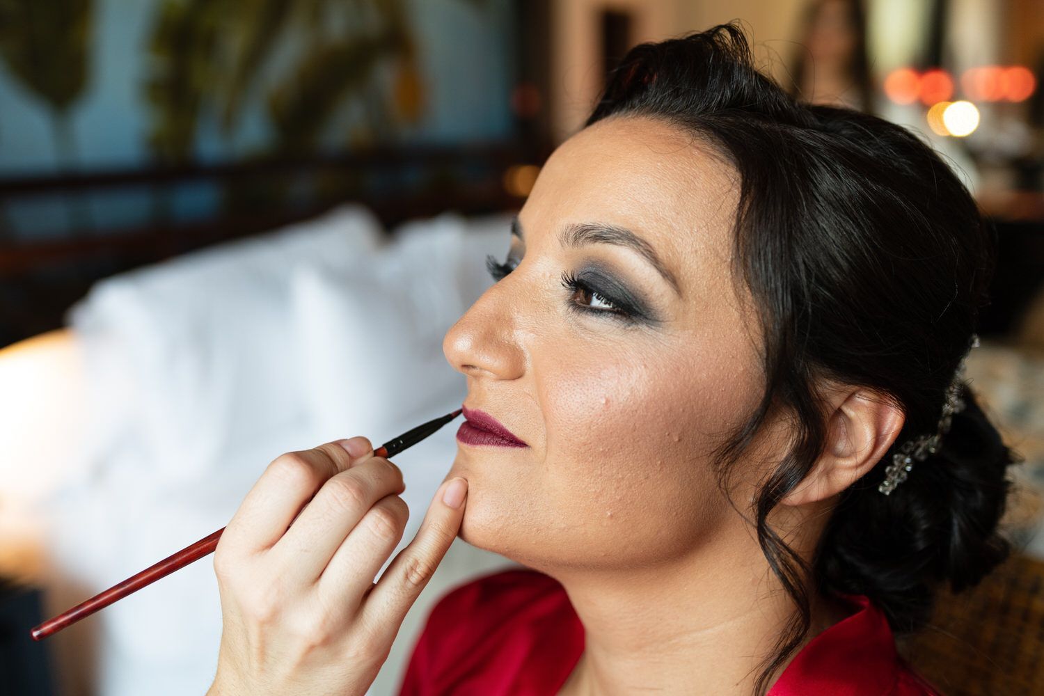 Mujer a la que le aplican lápiz labial, maquillaje de ojos oscuros, recogido, bata roja, artista sosteniendo un pincel