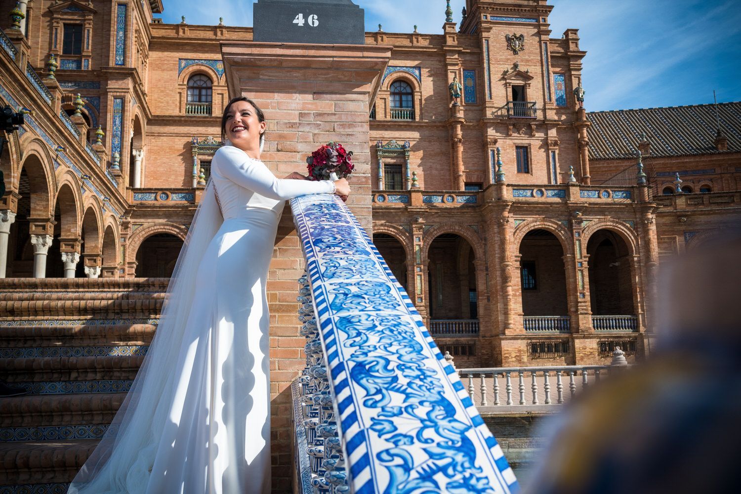 Una novia con un vestido blanco sonríe, sosteniendo un ramo, apoyada en una barandilla de azulejos azules y blancos 