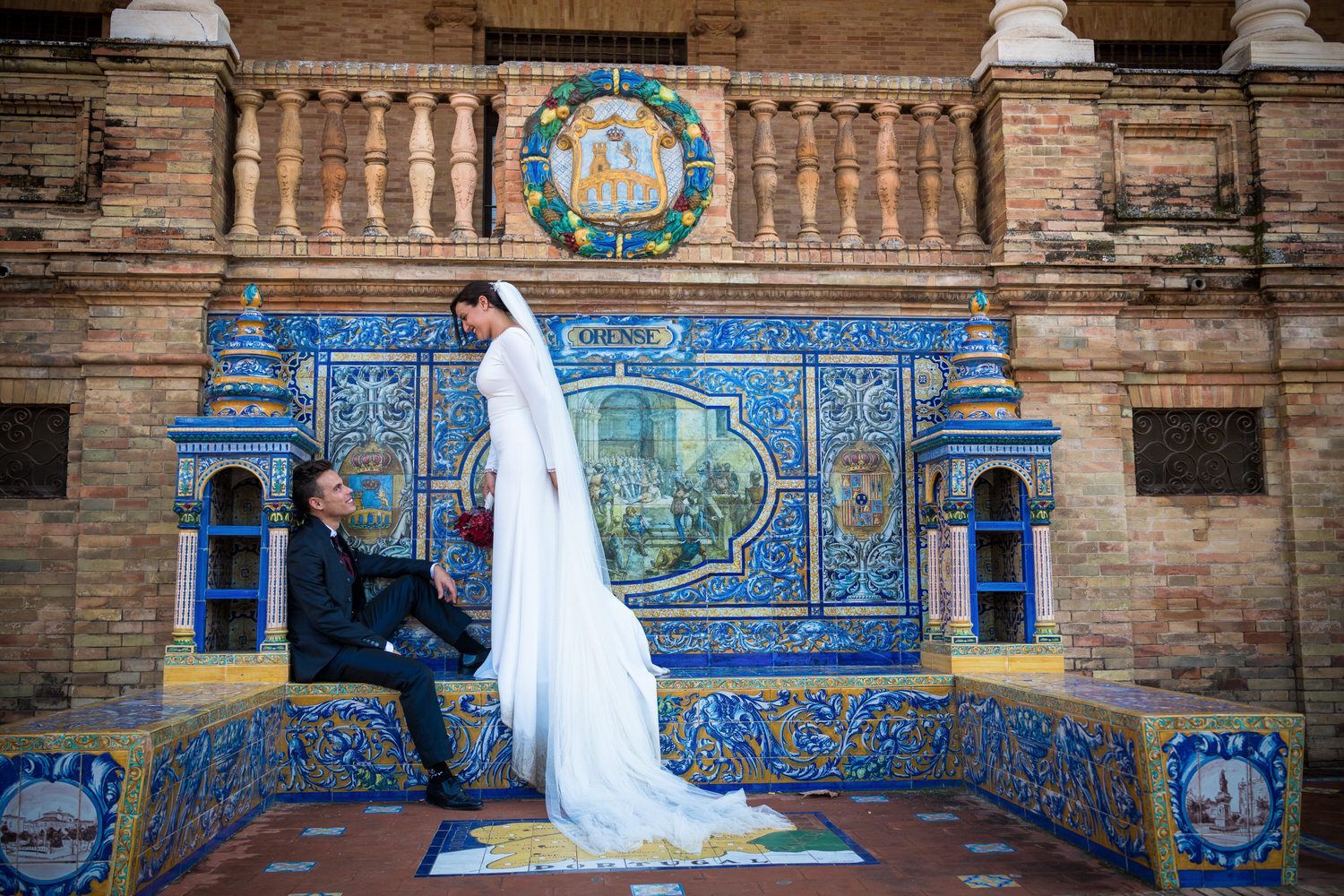 Los novios posan frente a una pared de azulejos azules ornamentados. Vestido de novia con cola larga. Atuendo formal.