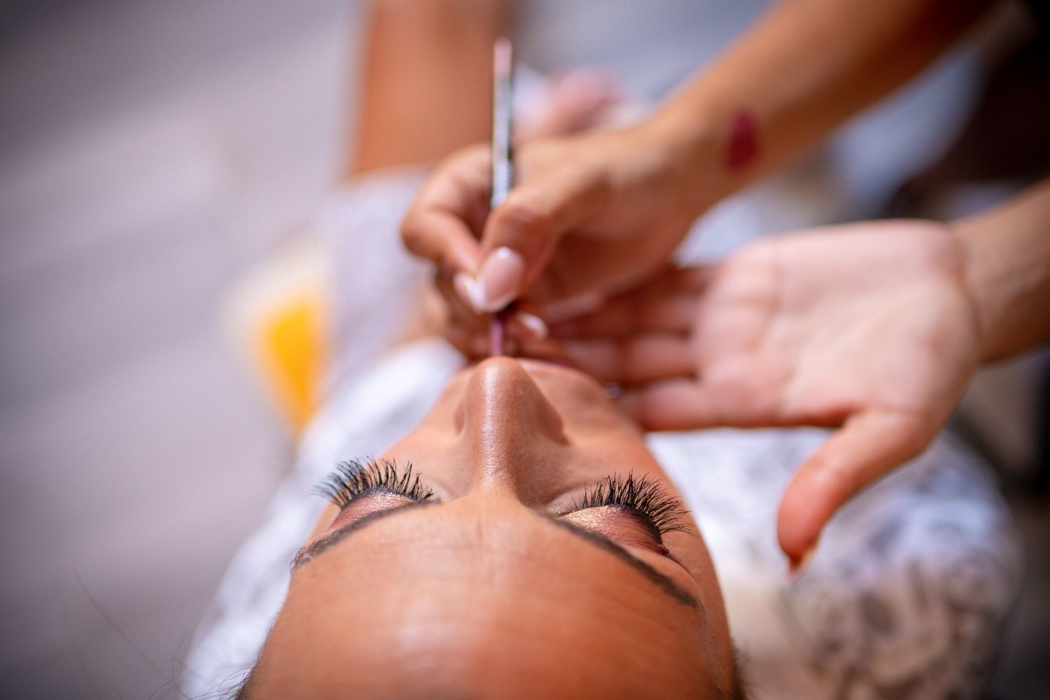 Persona maquillándose, primer plano. Ojos cerrados, pestañas largas. Maquilladora aplicando color de labios.