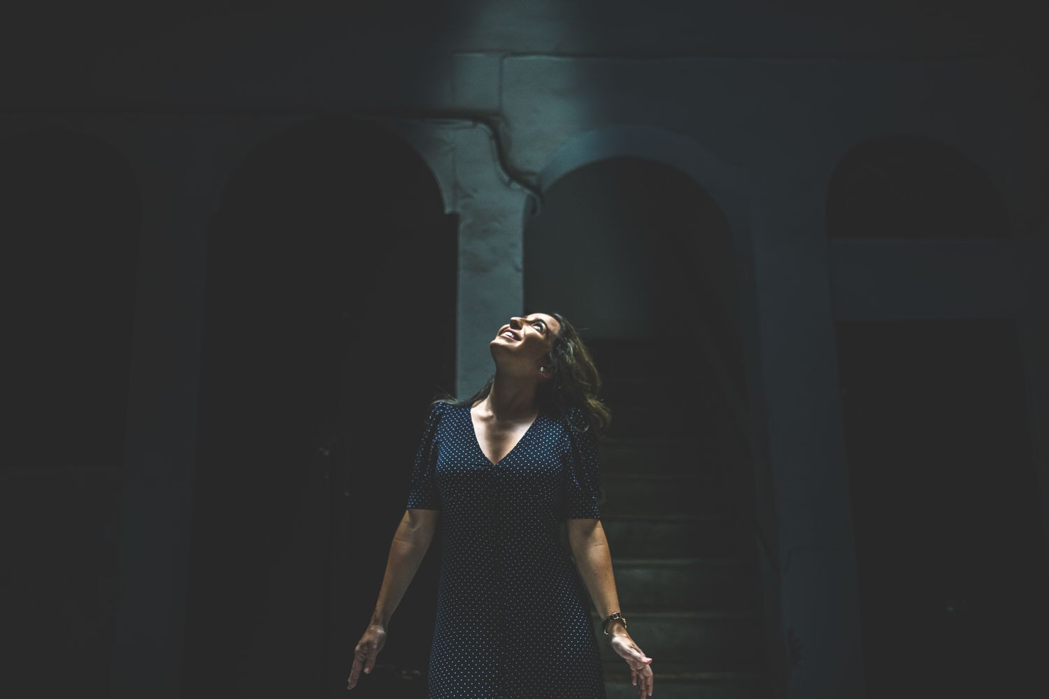 Una mujer en una habitación oscura mira hacia arriba con los brazos abiertos. La luz brilla desde arriba.
