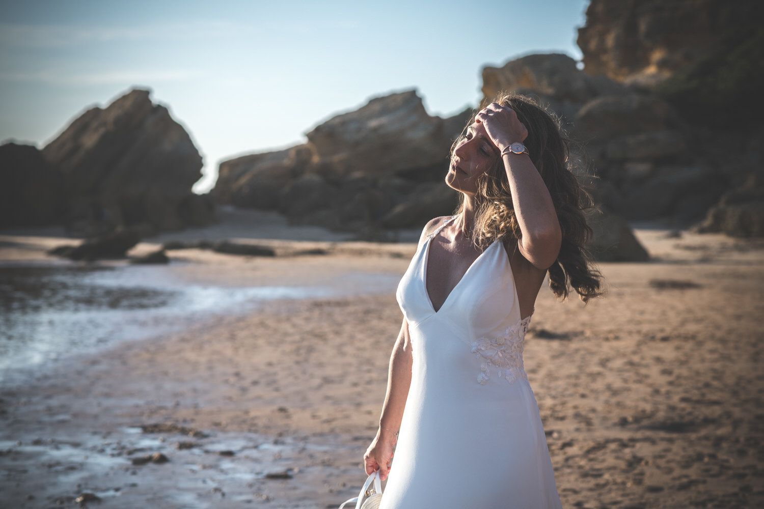 Novia con vestido blanco en una playa, tocándose el cabello, iluminada por el sol, fondo rocoso.