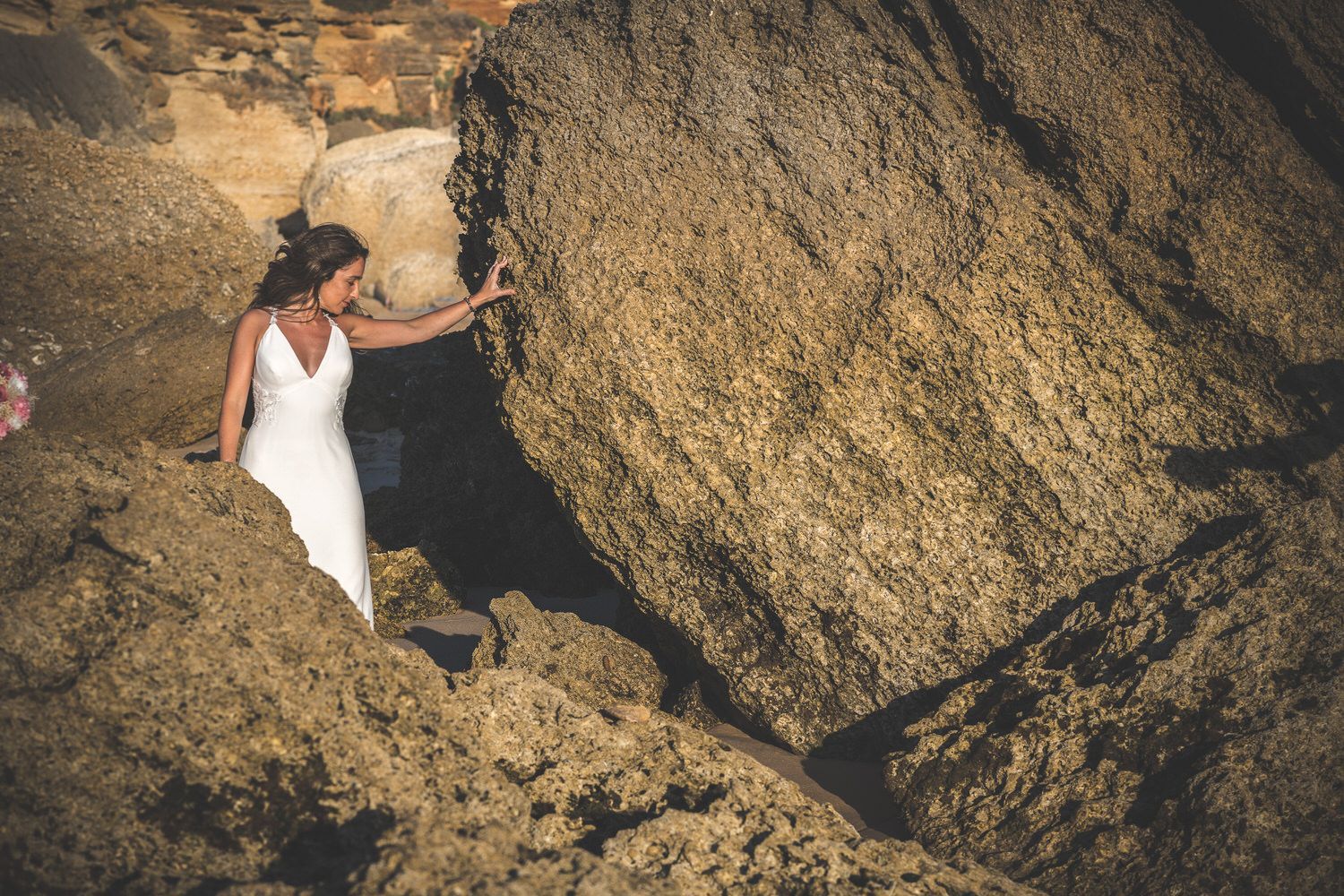 Una mujer vestida de blanco toca una gran formación rocosa texturizada cerca de la costa.