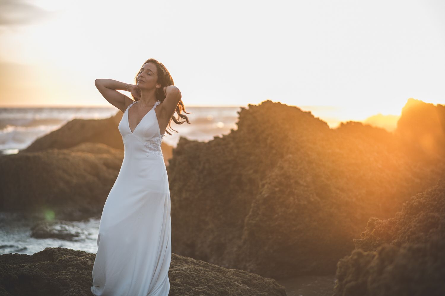 Mujer vestida de blanco con los brazos levantados, en la orilla rocosa al atardecer.