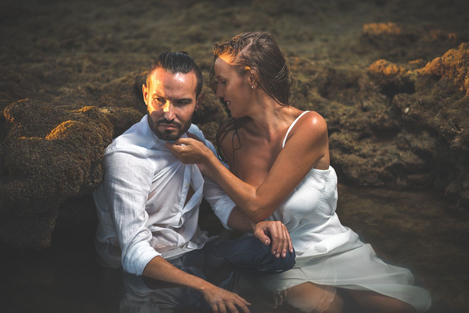 Pareja con ropa mojada, mujer acariciando la cara del hombre, sentados en el agua cerca de una orilla rocosa.