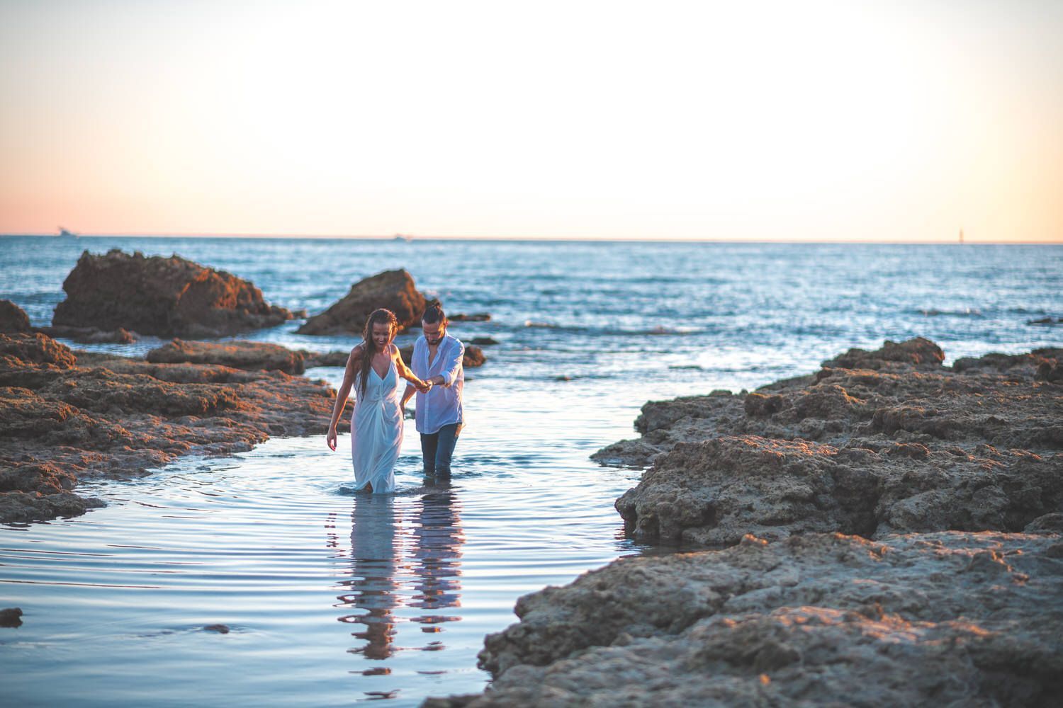 Pareja caminando en aguas poco profundas al atardecer, tomados de la mano cerca de la costa rocosa.