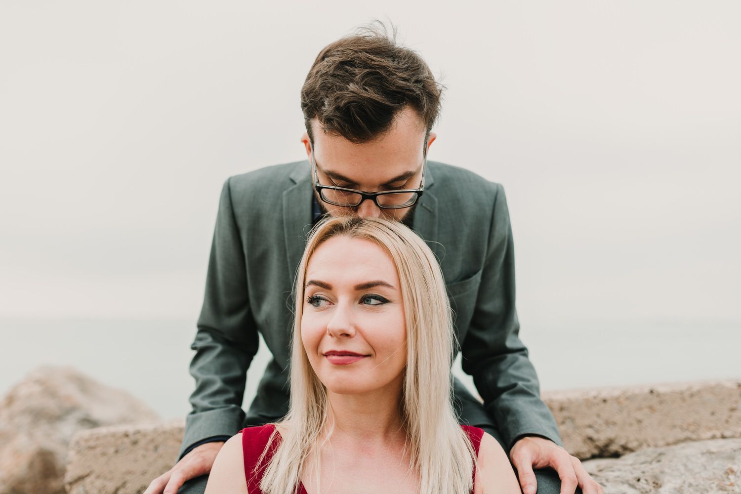 Un hombre besa a una mujer en la frente; están sentados al aire libre, cerca del agua. 
