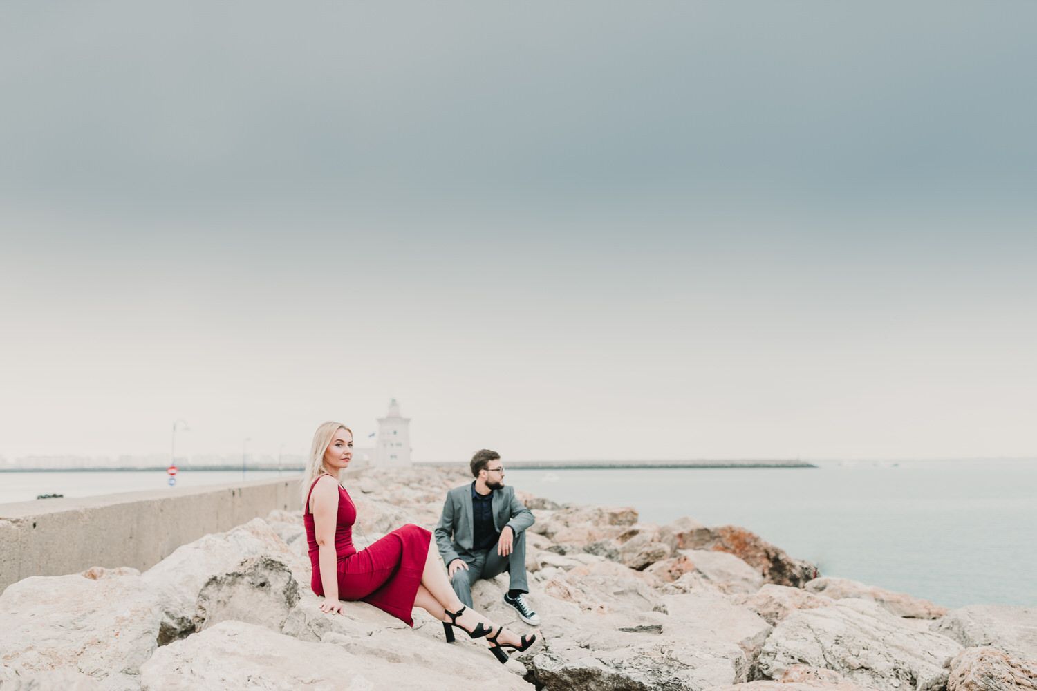 Una mujer vestida de rojo y un hombre con traje se sientan en las rocas y miran el mar.