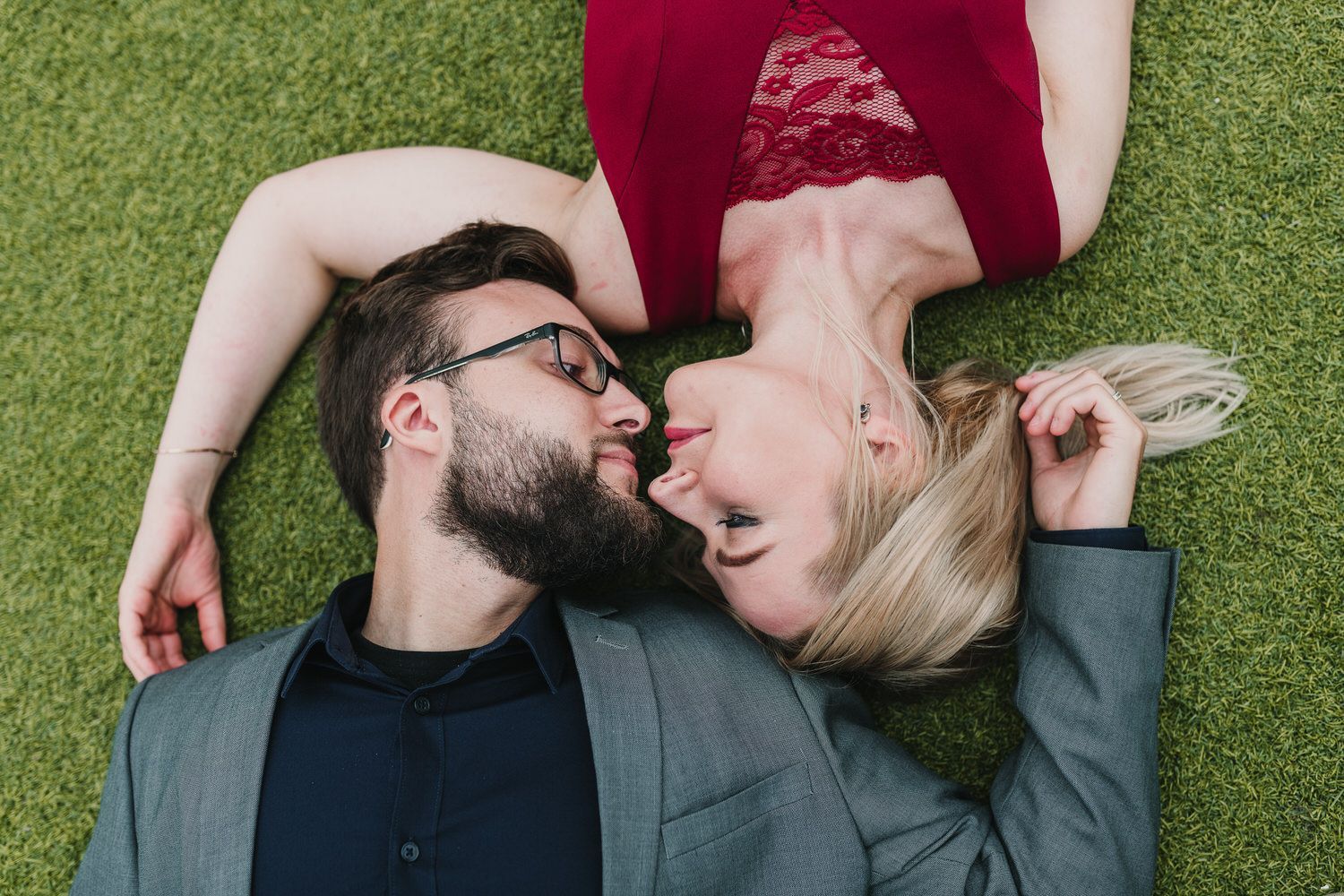 Pareja tumbada en el césped, caras juntas. La mujer lleva vestido rojo, el hombre lleva traje.